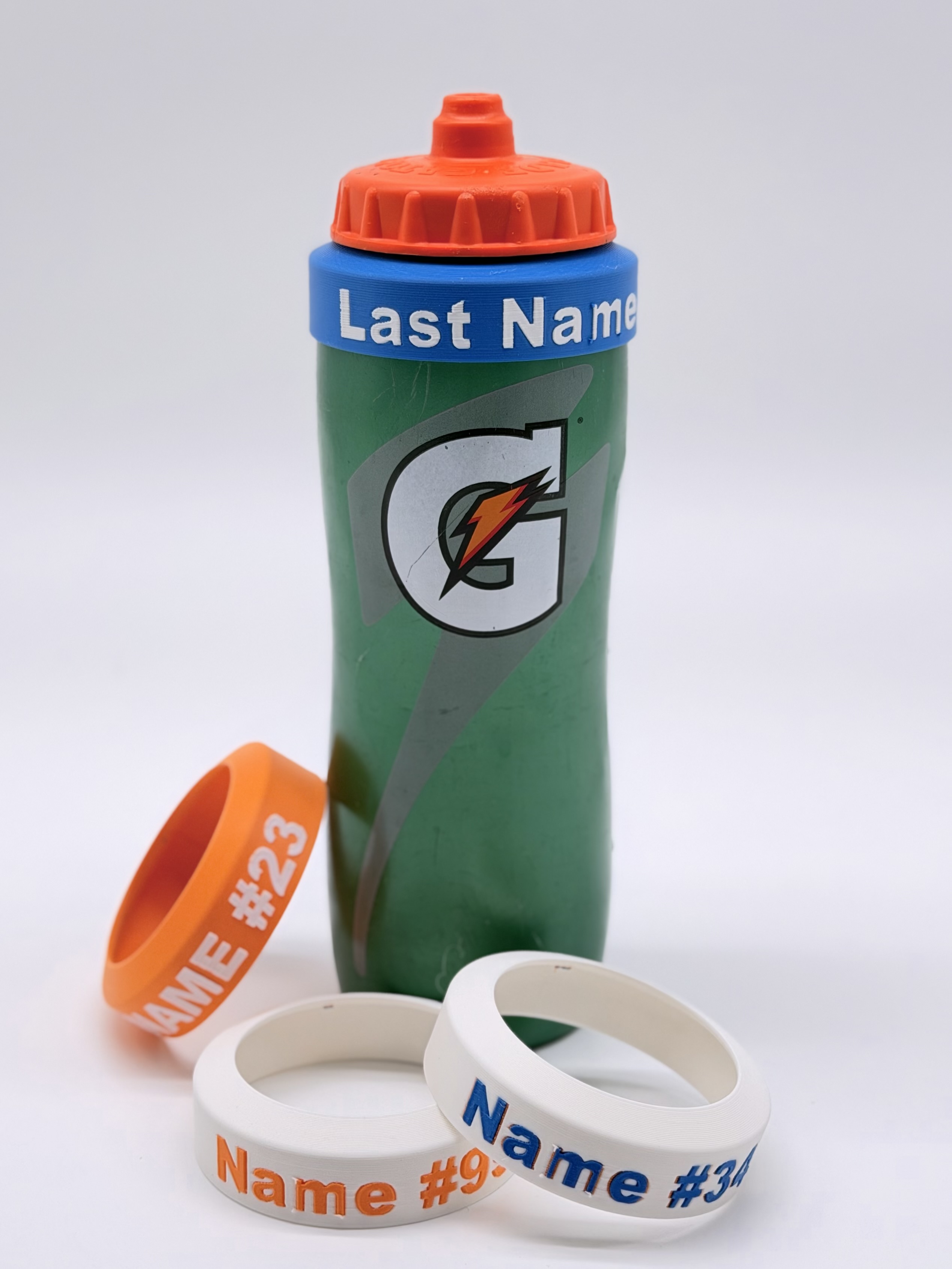 A green Gatorade sport bottle wearing a blue Last Name ring on its collar, with orange and white name-and-number rings resting beside it